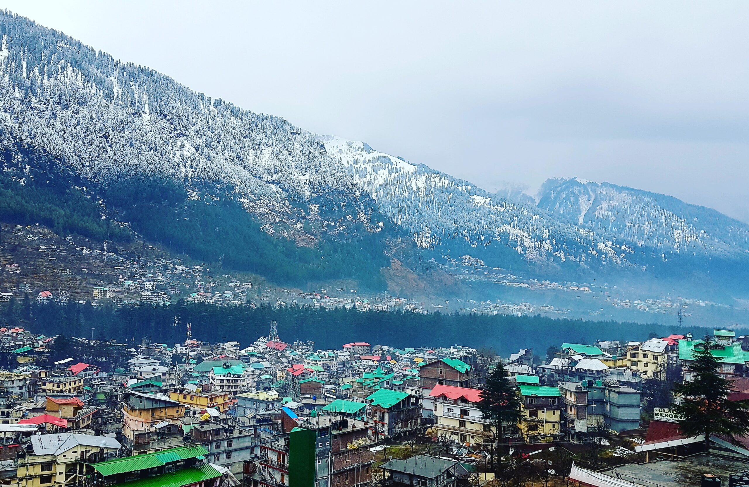 Scenic view of the town of Manali surrounded by pine forests and snow-covered mountains under a cloudy sky.